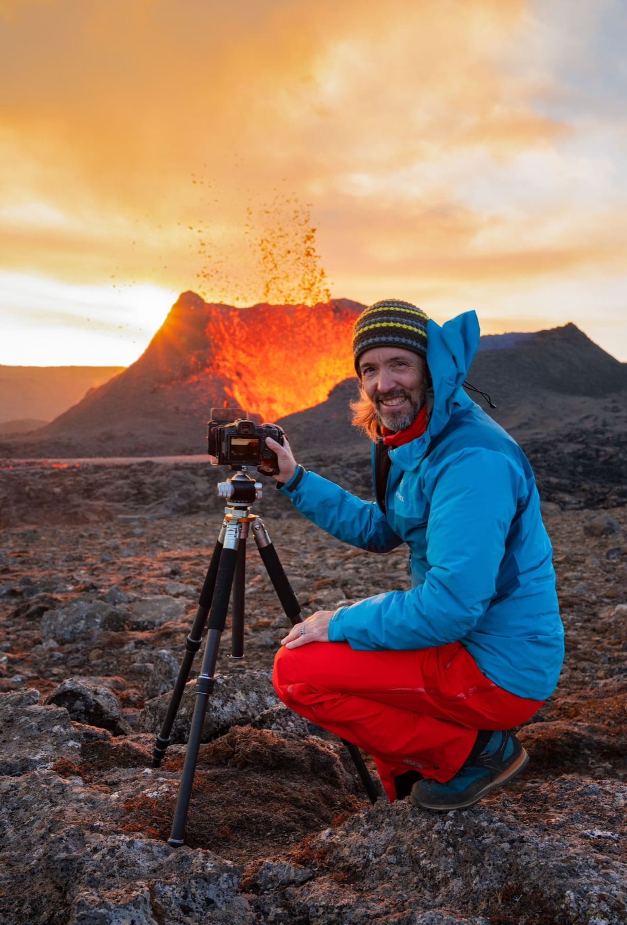 Dr. Oliver Schwenn crouching beside his tripod-mounted camera in front of an erupting volcano at sunset