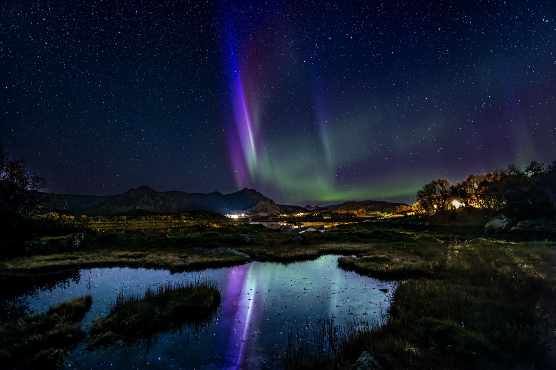 Violet and green aurora reflected in a still tarn under a starry sky